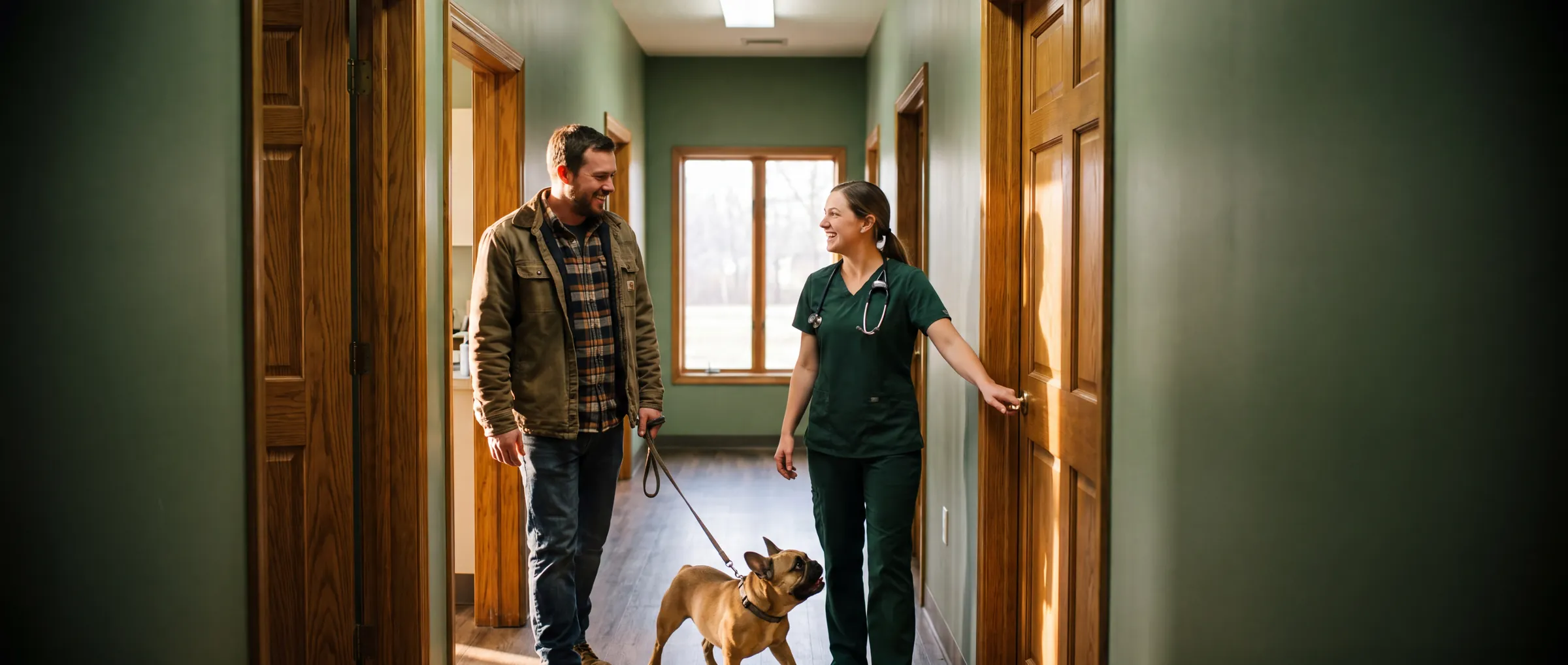 A veterinarian opens the exam-room door to greet the next patient, a French bulldog, and its owner